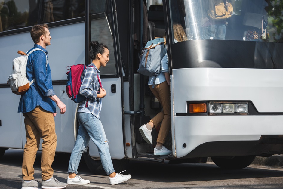 cheerful multiethnic friends with backpacks walking into travel bus at urban street